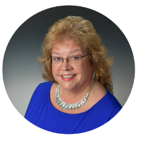 Headshot of Kentucky Farmers Bank VP/ Trust Op. Officer smiling, she is wearing a blue dress, glasses and a silver necklace against a grey background.